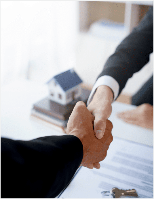 Two people shaking hands over a table with a model house in the background.