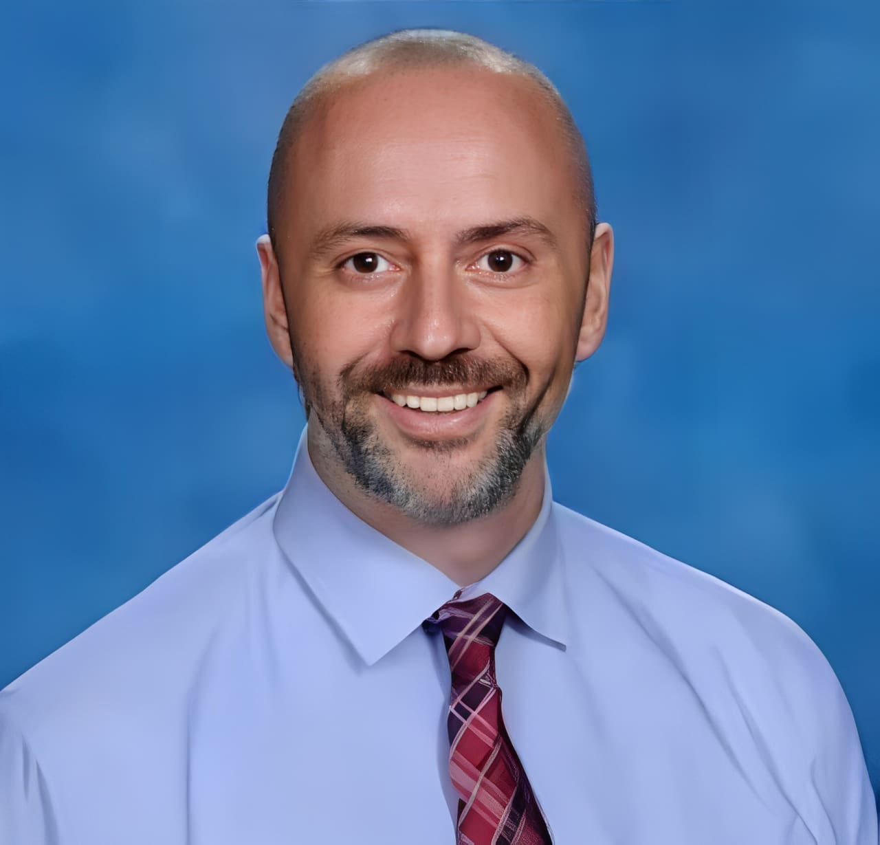 A man with a beard and tie in front of a blue background.